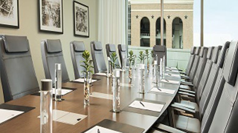 A modern conference room with a long table, chairs, water bottles, and notepads, facing large windows overlooking a building outside.