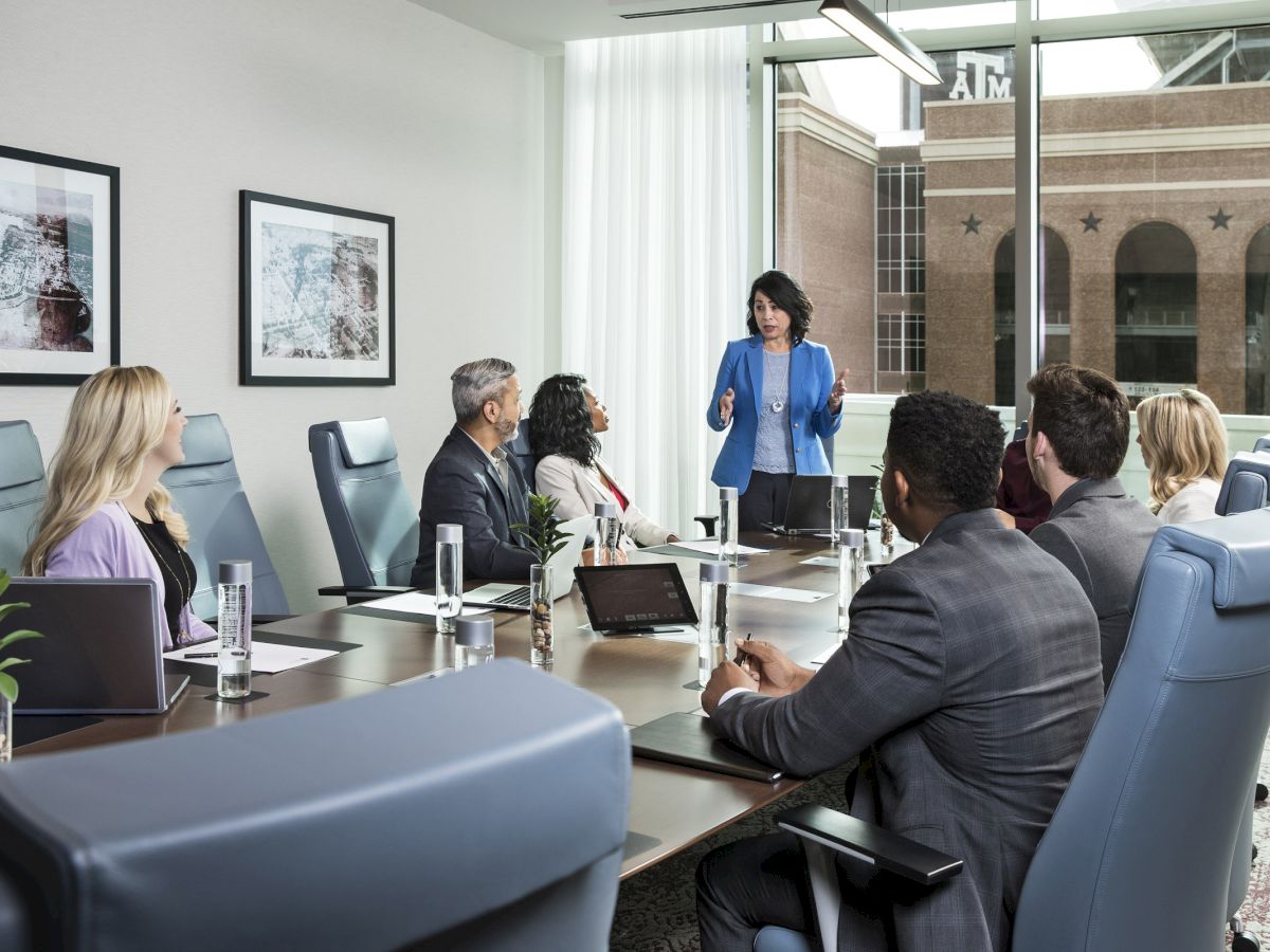 A business meeting with six people around a table, one person standing and presenting in a modern conference room with large windows and framed art.
