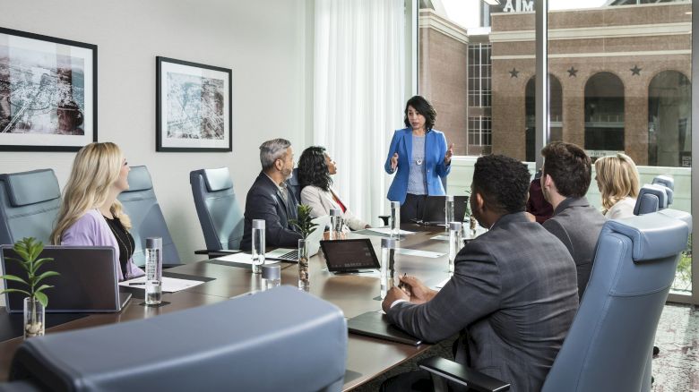 A business meeting with six people around a table, one person standing and presenting in a modern conference room with large windows and framed art.