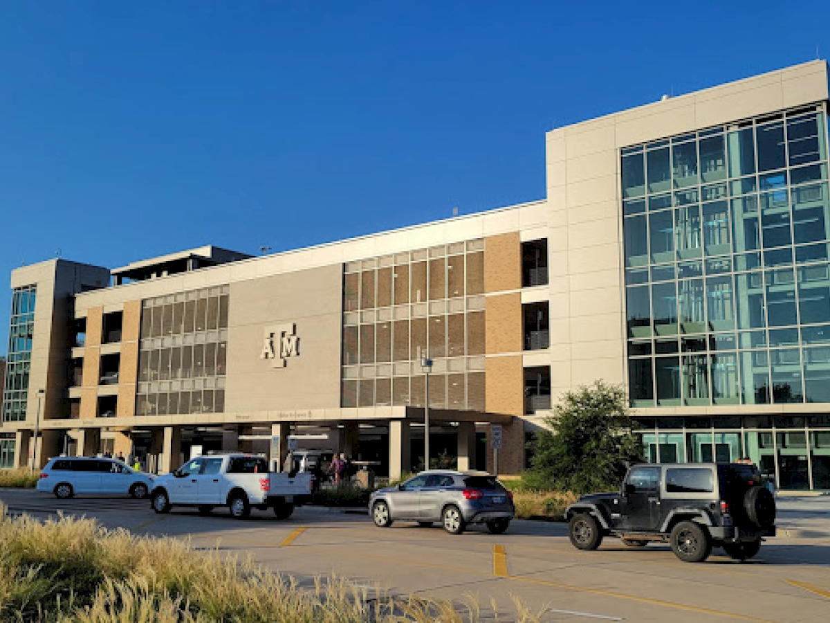 The image shows a modern multi-story building with a large "ATM" logo, a parking lot in the foreground, and several cars parked and moving around.