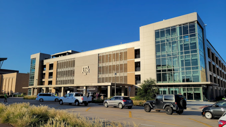 The image shows a modern multi-story building with a large "ATM" logo, a parking lot in the foreground, and several cars parked and moving around.
