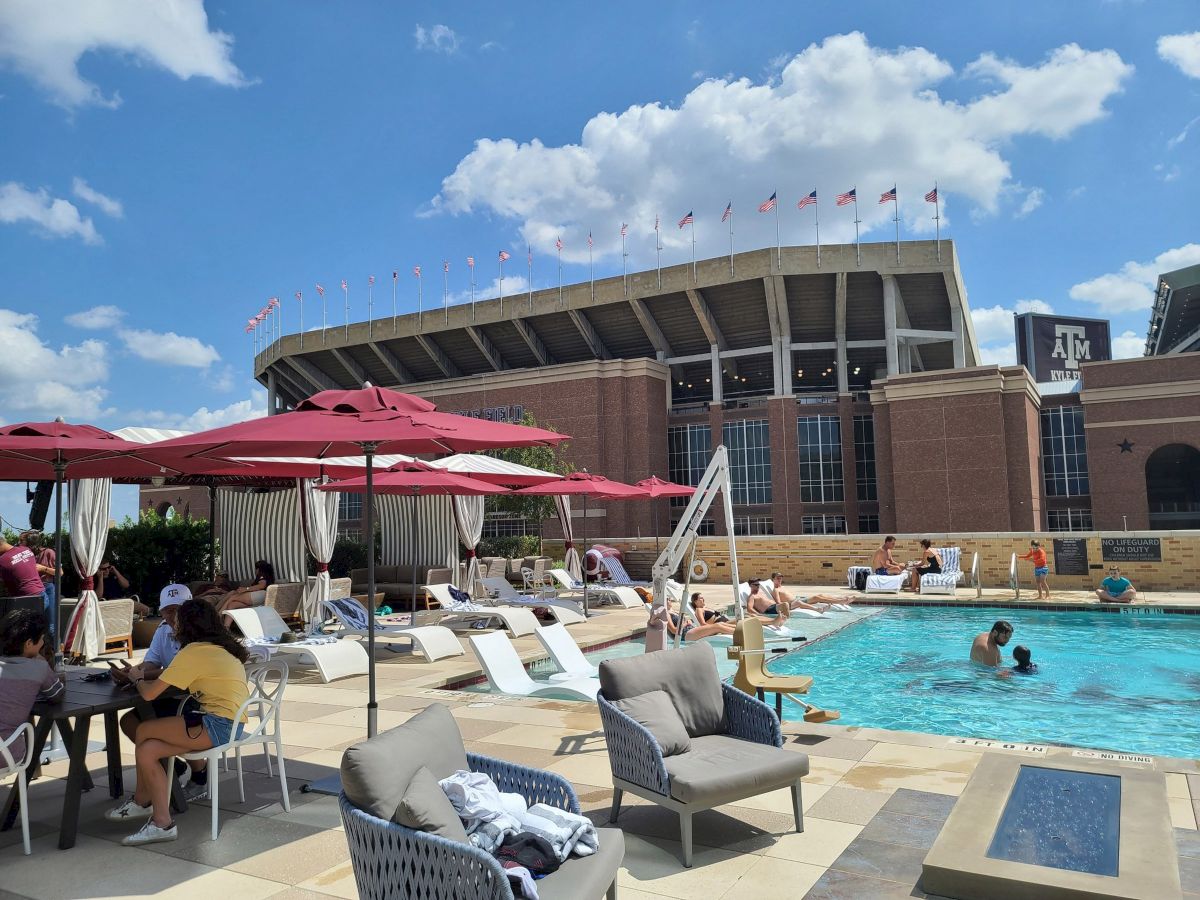 People relaxing by a pool with umbrellas and lounge chairs; a Kyle Field in the background under a partly cloudy sky.