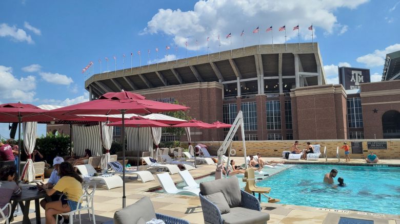 People relaxing by a pool with umbrellas and lounge chairs; a Kyle Field in the background under a partly cloudy sky.
