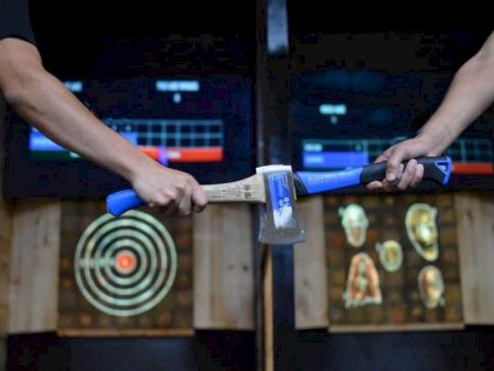 Two people holding axes with blue handles in front of targets in an indoor axe-throwing venue, with scoreboards visible in the background.
