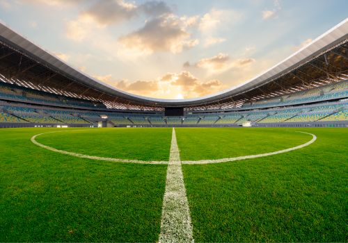 A pristine soccer stadium interior with a lush green pitch and a bold centerline, framed by empty stands under a cloudy sky.