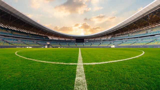 A pristine soccer stadium interior with a lush green pitch and a bold centerline, framed by empty stands under a cloudy sky.