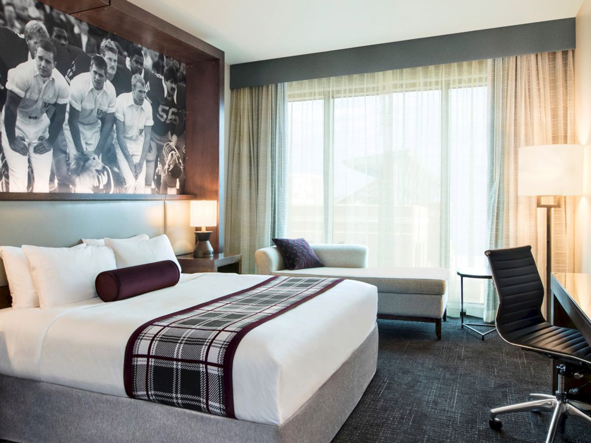 Photo of modern hotel room with a tide white bed with maroon throw, desk and chair, settee, and black and white photo of Aggie yell leaders