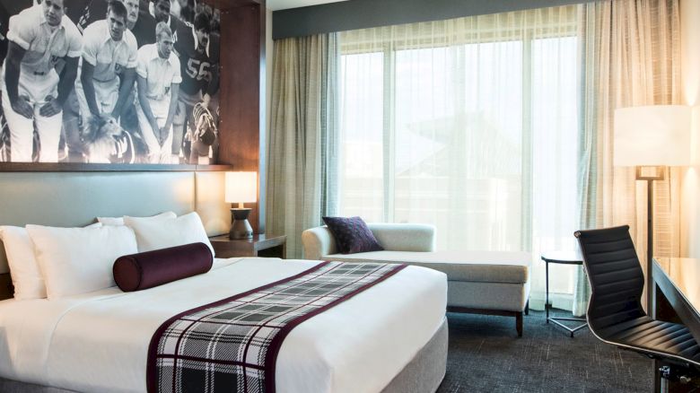 Photo of modern hotel room with a tide white bed with maroon throw, desk and chair, settee, and black and white photo of Aggie yell leaders
