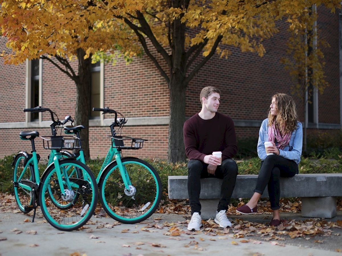 Veo Bikes parked beside a bench on campus with a man and woman holding a coffee and talking to one another