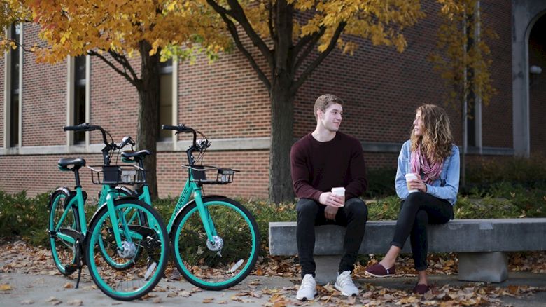 Veo Bikes parked beside a bench on campus with a man and woman holding a coffee and talking to one another