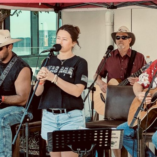 A band performs live on an outdoor stage: guitarist, vocalists, and a drummer under a red canopy, wearing casual summer outfits.