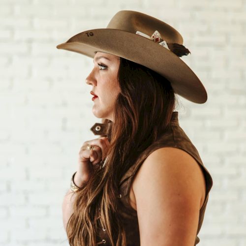 A woman in a wide-brim cowboy hat stands in profile, looking off to the side against a light background.