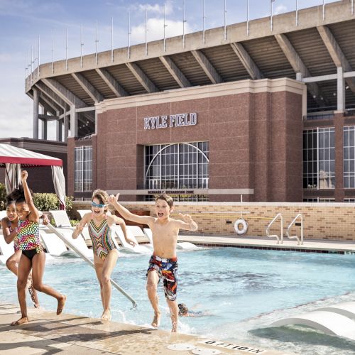 Kids splash and run toward the pool at a resort with lounge chairs, umbrellas, and a stadium-style building in the background.