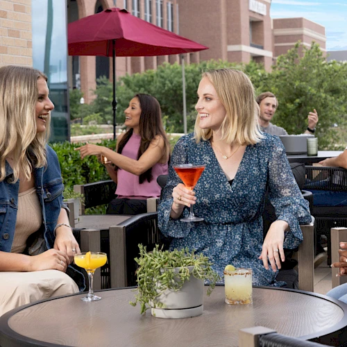 Four friends chat and sip colorful cocktails on a sunny outdoor patio, with umbrellas, brick walls, and greenery in the background.