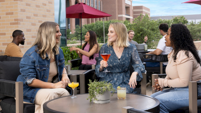 Four friends chat and sip colorful cocktails on a sunny outdoor patio, with umbrellas, brick walls, and greenery in the background.