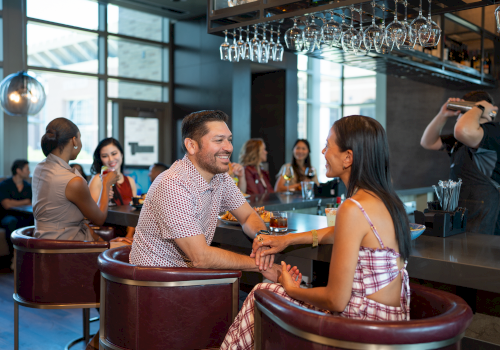 A couple chats warmly at a bar while others sip and mingle in the background, capturing a lively, social evening scene.