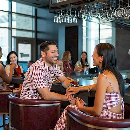 A couple chats warmly at a bar while others sip and mingle in the background, capturing a lively, social evening scene.