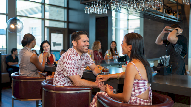 A couple chats warmly at a bar while others sip and mingle in the background, capturing a lively, social evening scene.