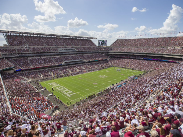 A packed American football stadium with fans in the stands and a live game on the green field under a cloudy sky.