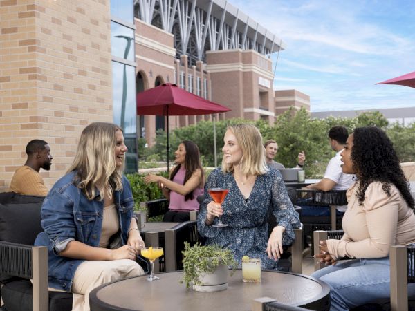 A group of friends lounges at an outdoor patio table with drinks, chatting and smiling under pink umbrellas in front of a brick building.