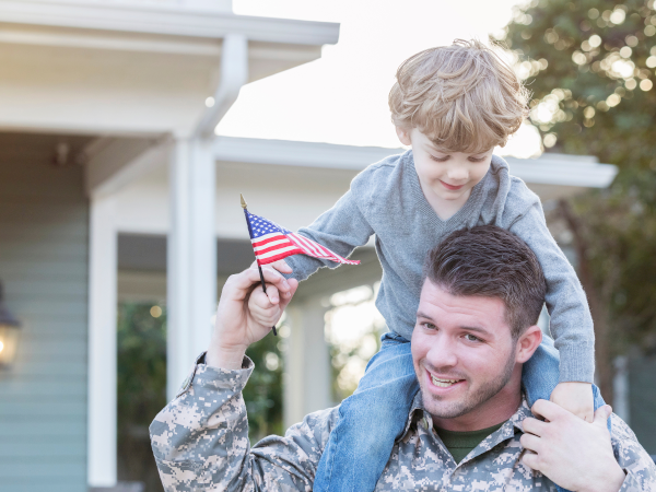 A soldier in camouflage holds a little boy who waves a small American flag outdoors near a house.