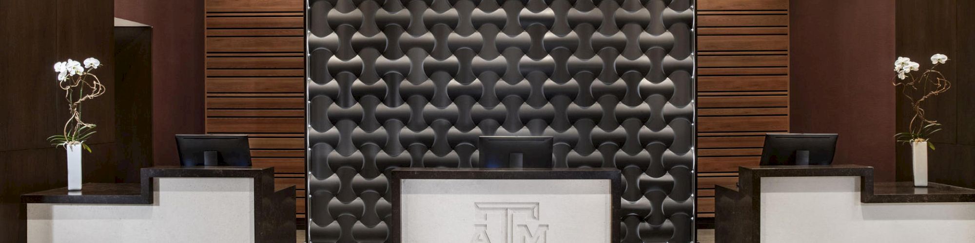 A modern reception area with three desks, a decorative wall with a black pattern, wooden accents, two pendant lights, and white orchid plants.