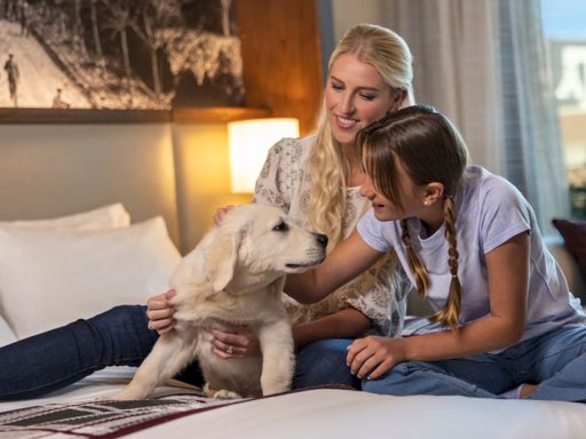 A woman and a girl are sitting on a bed petting a fluffy puppy, all looking happy in a cozy room.