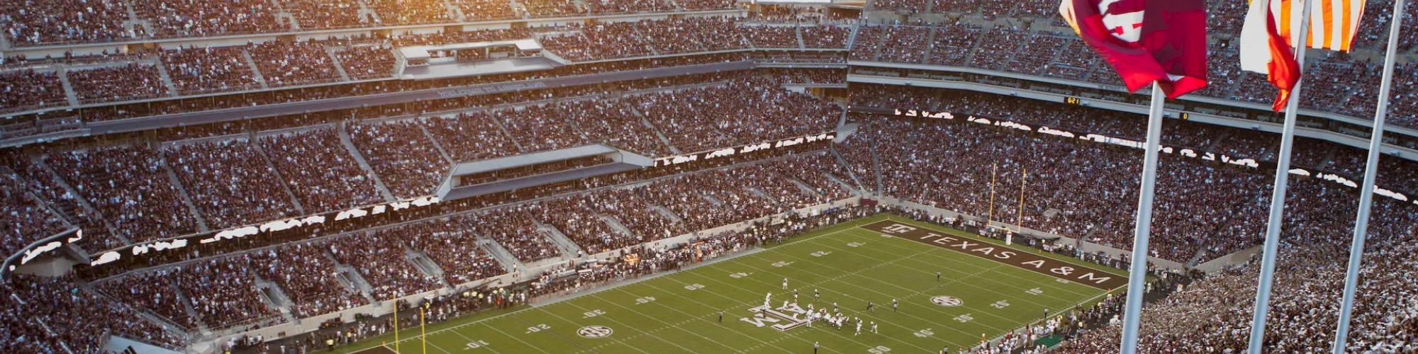 A packed football stadium with "Texas A&M" on the field, flags waving, and the stands filled with spectators enjoying the game at sunset.