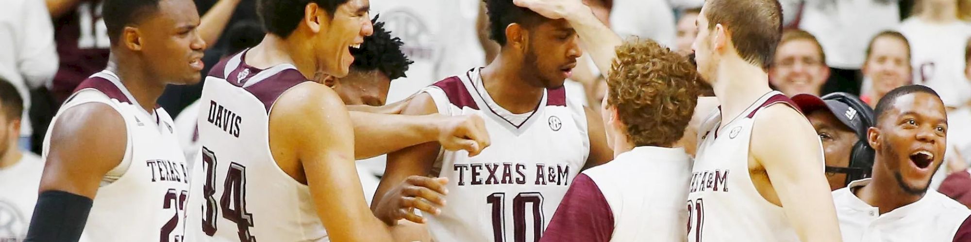 A basketball team in white jerseys with maroon accents, labeled "Texas A&M," celebrates on the court surrounded by fans in white.