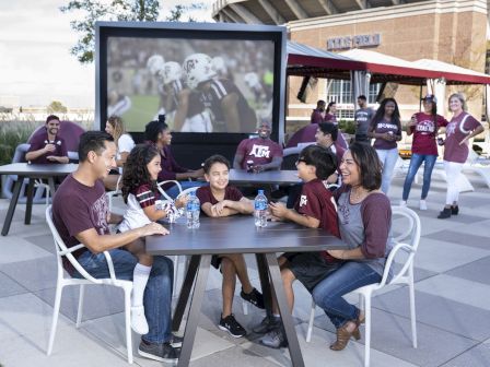 A group of people in Texas A&M maroon shirts sit outside, watching a game on a big screen in a stadium event area.