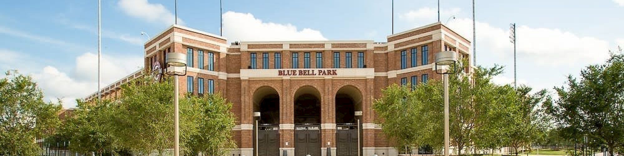 The image shows the entrance to Blue Bell Park, a sports stadium surrounded by trees and landscaping under a partly cloudy sky.