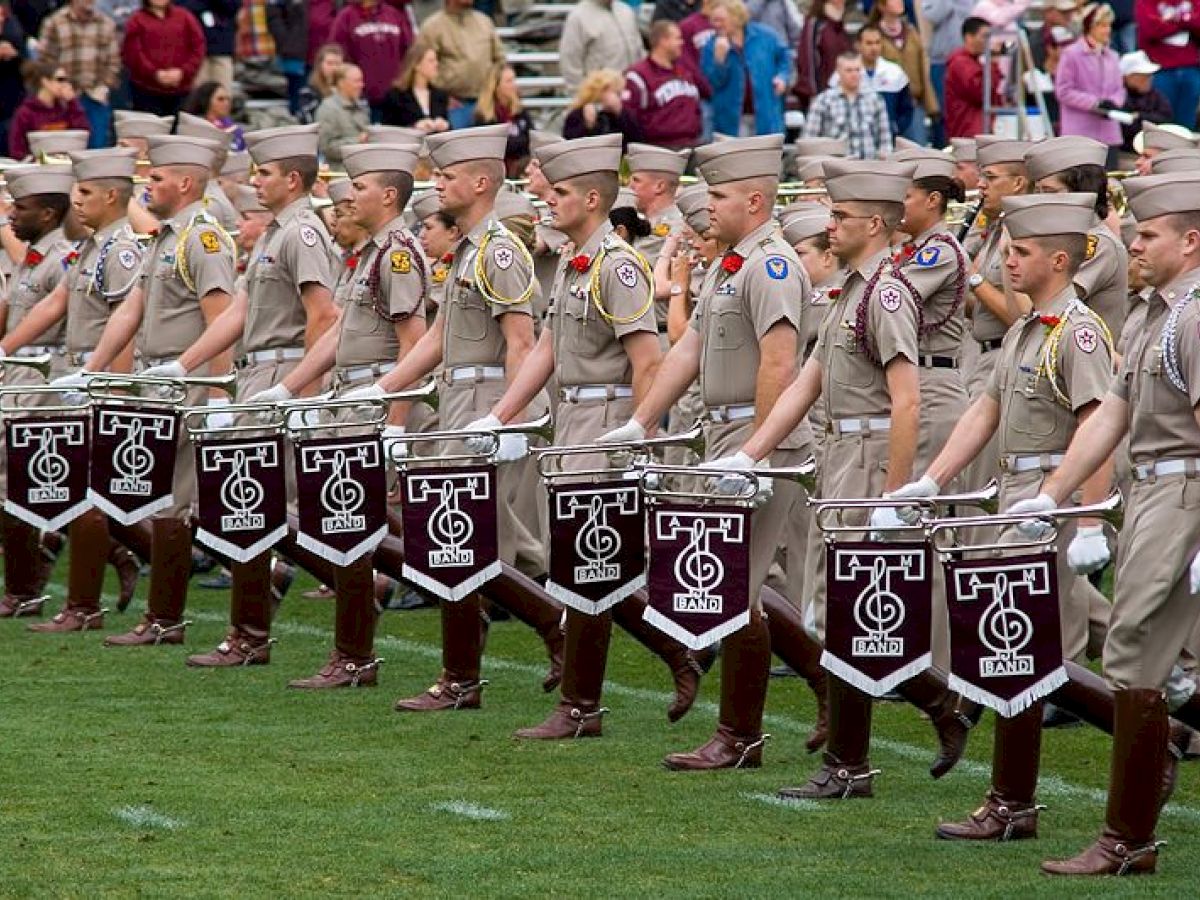 A group of uniformed individuals is marching on a field, each carrying a snare drum with the letter T and a symbol on the front, while a crowd watches.