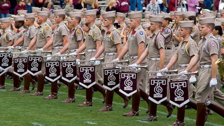 A group of uniformed individuals is marching on a field, each carrying a snare drum with the letter T and a symbol on the front, while a crowd watches.