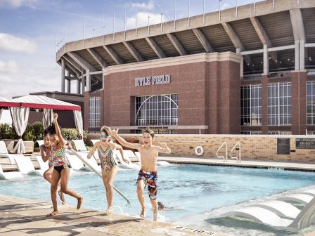 Four children are joyfully running along the edge of a pool at an outdoor setting with a stadium in the background.