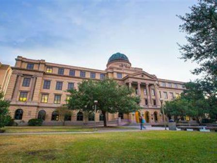 The image shows a grand, historic building with a dome and columns, surrounded by green trees and an open lawn, under a blue sky.