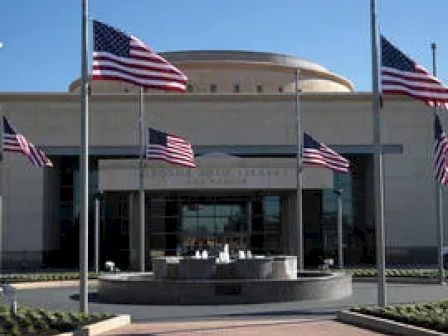 The image shows an entrance of a building with a fountain in front, surrounded by multiple American flags on poles, and a domed structure in the background.