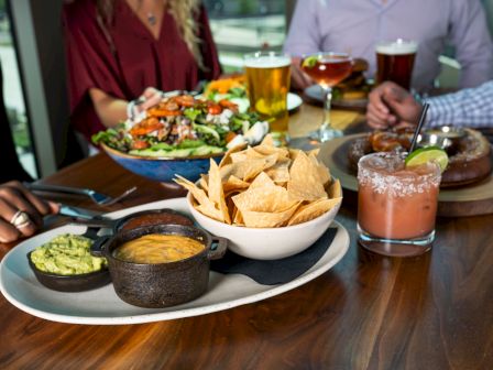 The image shows a table with people enjoying a meal, featuring a bowl of tortilla chips with dips, a salad, drinks, and an appetizer on a wooden table.