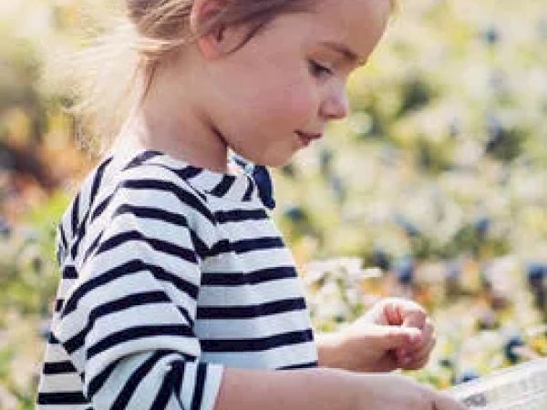 A young child with a striped shirt is outdoors in a field, holding a clear container. The background is sunny with greenery and flowers.
