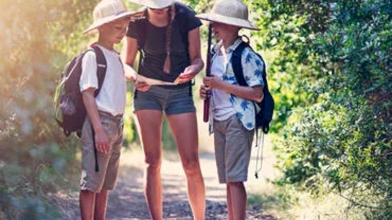A woman and two children wearing hats and backpacks are looking at a map while standing on a forest trail, surrounded by greenery and sunlight.