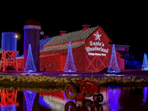 A festively lit barn with "Santa's Wonderland" signage, surrounded by illuminated Christmas trees and a small red tractor by a reflective pond.
