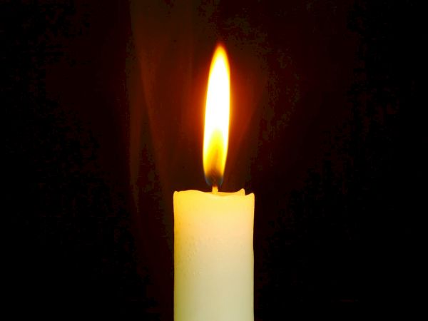 The image shows a single white candle burning with a bright flame against a dark background.