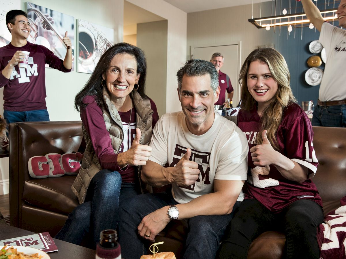A happy group wearing Texas A&M gear poses on a couch with thumbs up in a cozy game-day living room.