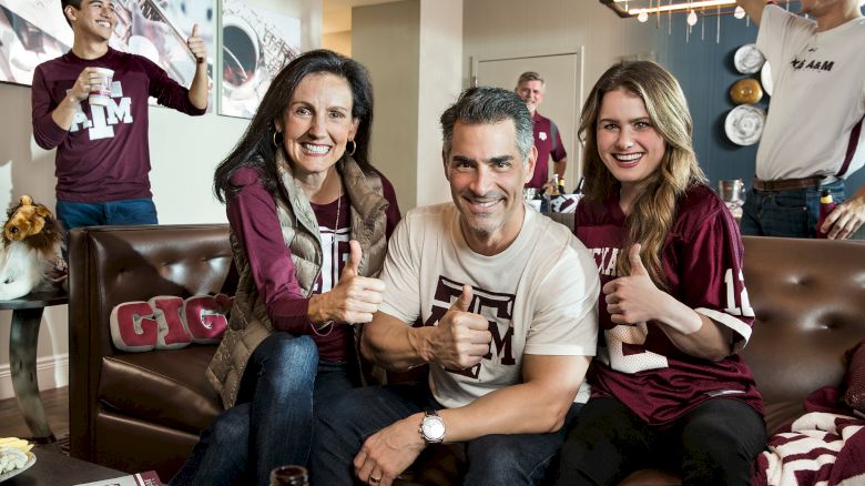 A happy group wearing Texas A&M gear poses on a couch with thumbs up in a cozy game-day living room.