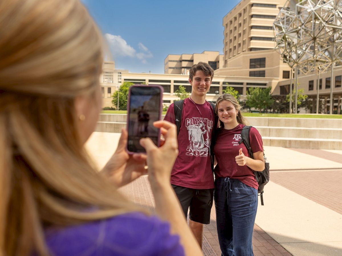 A person is taking a photo of two people standing side by side, with a university campus in the background and a unique spherical structure visible.