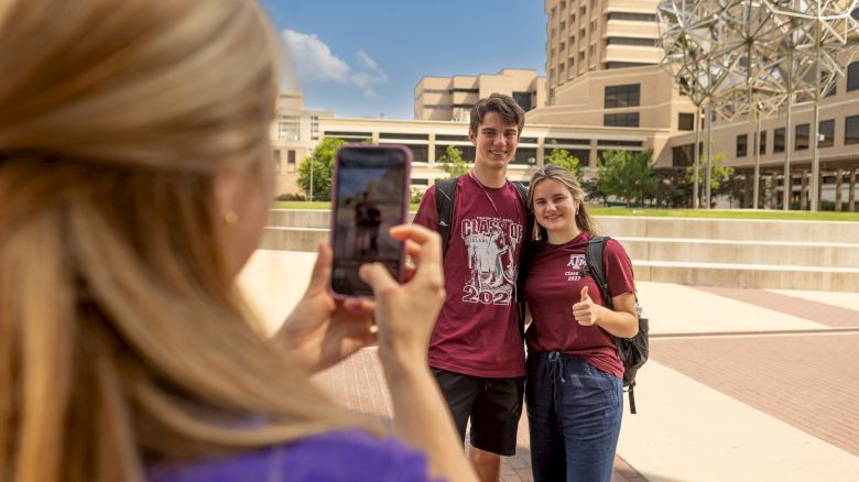 A person is taking a photo of two people standing side by side, with a university campus in the background and a unique spherical structure visible.