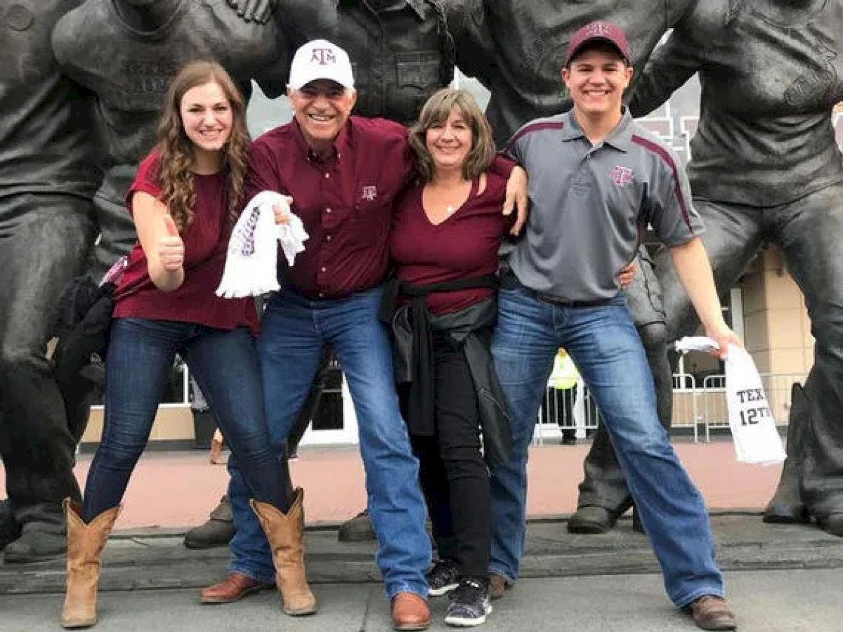A group of four people, two men and two women, are happily posing in front of a statue, wearing maroon and gray attire, and holding white towels.