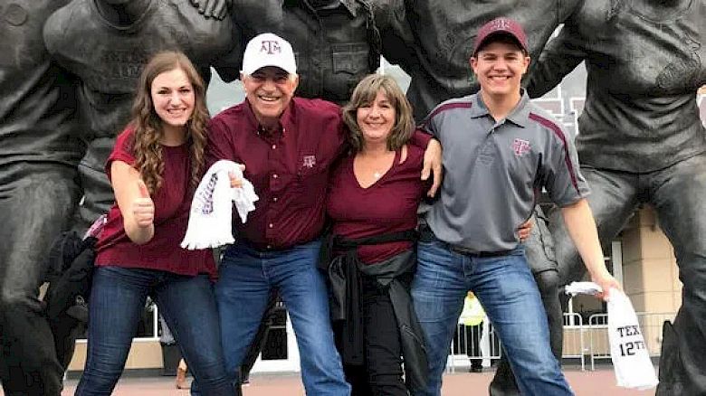 A group of four people, two men and two women, are happily posing in front of a statue, wearing maroon and gray attire, and holding white towels.