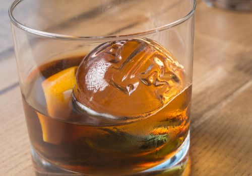A glass of whiskey with a spherical ice cube and a twist of lemon, placed on a wooden surface.
