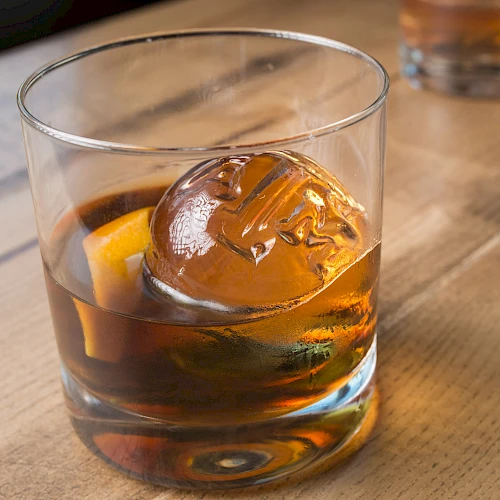 A glass of whiskey with a spherical ice cube and a twist of lemon, placed on a wooden surface.