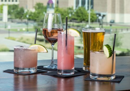 The image shows five different drinks, including cocktails, a beer, and a wine glass, placed on a table with a large brick building in the background.